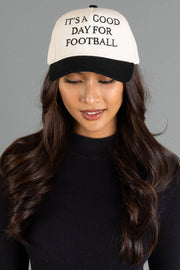 A woman with long brown hair, wearing a black top and the Panache Apparel Collection’s It's A Good Day For Football Two-Toned Trucker Hat—featuring a white crown, black brim, and adjustable snapback—is looking down with a slight smile.