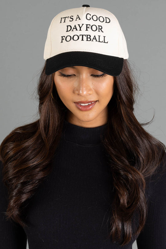 A woman with long brown hair, wearing a black top and the Panache Apparel Collection’s It's A Good Day For Football Two-Toned Trucker Hat—featuring a white crown, black brim, and adjustable snapback—is looking down with a slight smile.