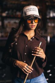 A woman wearing the Panache Apparel Collection World’s Best Ex-Girlfriend Tan & Camo Two-Toned Trucker Hat, large sunglasses, a brown button-up shirt, and jeans holds a pool cue in a dimly lit bar or billiards room.