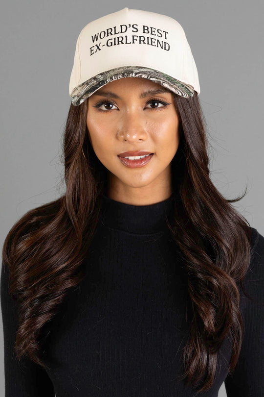 A woman with long dark hair is wearing the Panache Apparel Collection “World’s Best Ex-Girlfriend” Tan & Camo Two-Toned Trucker Hat and a black sweater, posing against a plain gray background.