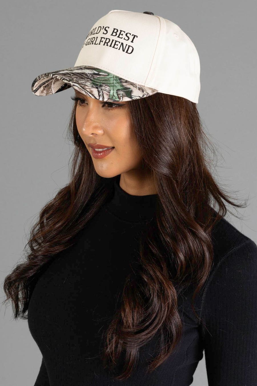 Woman with long brown hair wearing a Panache Apparel Collection World’s Best Ex-Girlfriend Tan & Camo Two-Toned Trucker Hat and a black long-sleeve top, posing against a plain gray background.