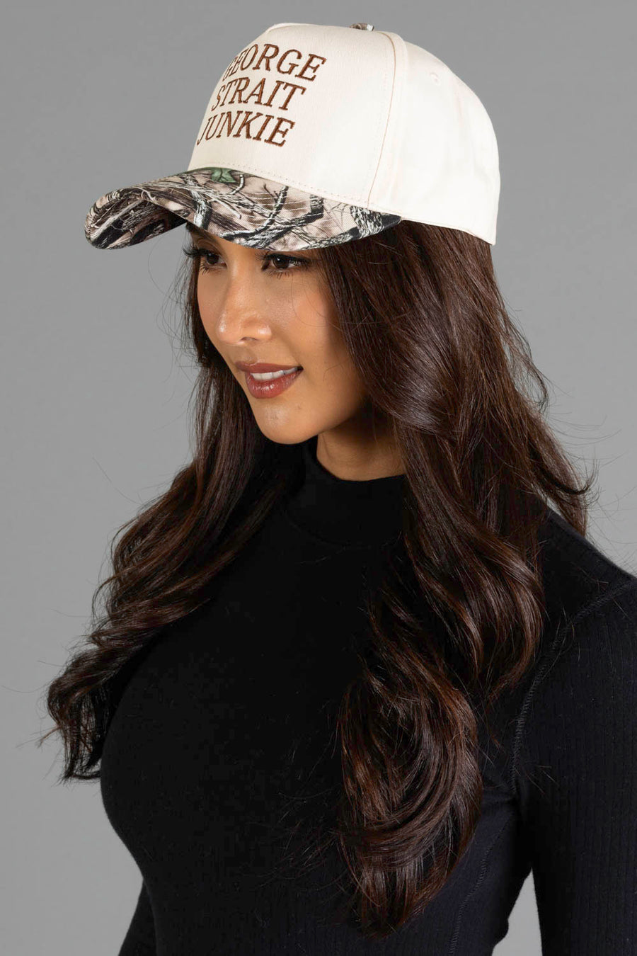 Woman with long brown hair wearing the Panache Apparel Collection’s George Strait Junkie Tan & Camo Two-Toned Trucker Hat and a black top, smiling and looking to the side against a gray background.