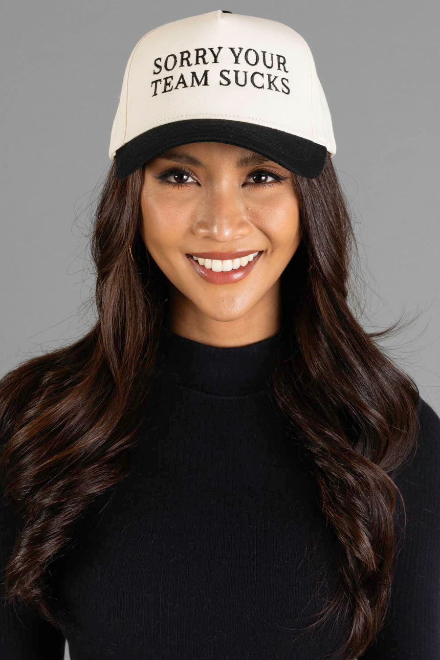 A smiling woman with long dark hair wears a black sweater and the Panache Apparel Collection Sorry Your Team Sucks Two-Toned Trucker Hat—an adjustable snapback with white embroidery—set against a plain gray background.
