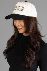 A woman with long brown hair, dressed in a black turtleneck, smiles and looks down while wearing the Panache Apparel Collection's Sorry Your Team Sucks Two-Toned Trucker Hat.