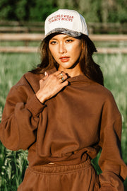 A woman in a brown sweatshirt and sweatpants stands outdoors, wearing the Panache Apparel Collection Hot Girls Wear Cowboy Boots Tan & Camo Two-Toned Trucker Hat—featuring a white front, camouflage brim, and “HOT GIRLS WEAR COWBOY BOOTS” embroidered in red—while touching her neck with one hand.