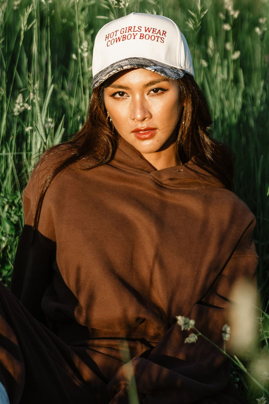 A woman with long brown hair wears a brown hoodie and the Panache Apparel Collection Hot Girls Wear Cowboy Boots Tan & Camo Two-Toned Trucker Hat while sitting in tall grass, sunlight illuminating her face.