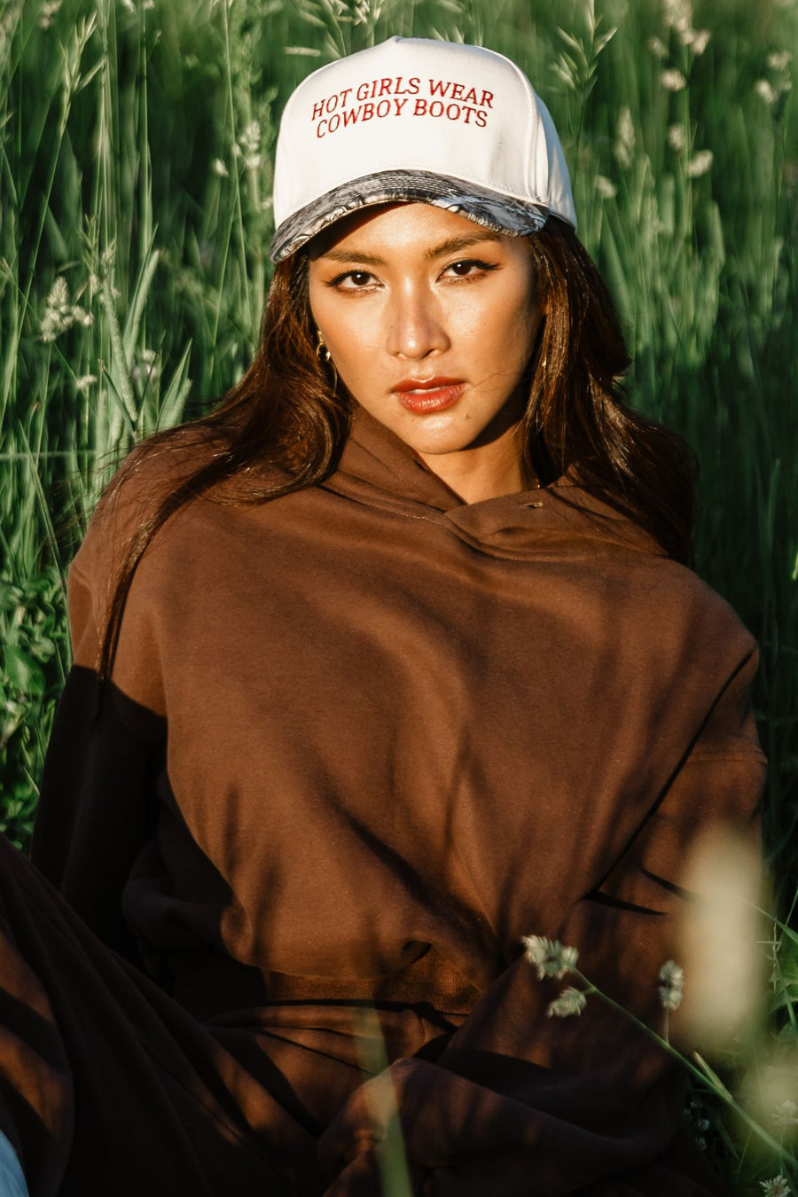 A woman with long brown hair wears a brown hoodie and the Panache Apparel Collection Hot Girls Wear Cowboy Boots Tan & Camo Two-Toned Trucker Hat while sitting in tall grass, sunlight illuminating her face.
