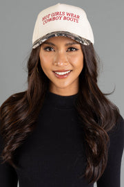 A woman with long wavy hair smiles in front of a gray background, wearing a black ribbed top and the Panache Apparel Collection Hot Girls Wear Cowboy Boots Tan & Camo Two-Toned Trucker Hat, which features red lettering on the front.