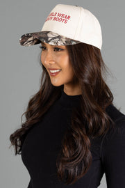 A woman in a black top wears the Panache Apparel Collection “Hot Girls Wear Cowboy Boots” tan and camo two-toned trucker hat, smiling and looking to the side against a gray background.