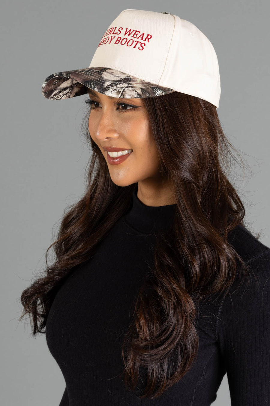 A woman in a black top wears the Panache Apparel Collection “Hot Girls Wear Cowboy Boots” tan and camo two-toned trucker hat, smiling and looking to the side against a gray background.