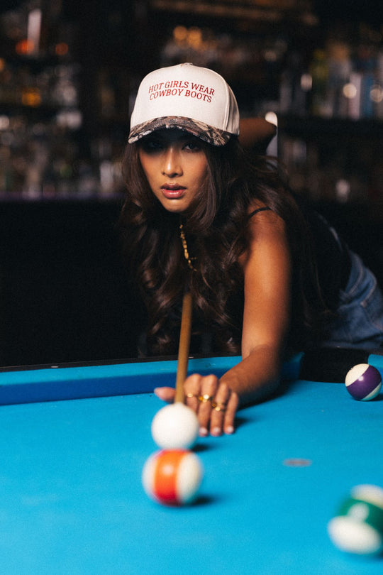 A woman sporting the Panache Apparel Collection’s Hot Girls Wear Cowboy Boots Tan & Camo Two-Toned Trucker Hat and cowboy boots leans over a blue pool table, aiming her cue for a shot.