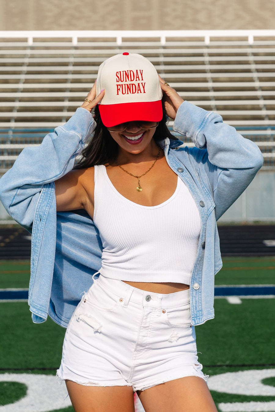 A woman stands on a sports field, smiling as she adjusts her Panache Apparel Collection Sunday Funday Two-Toned Trucker Hat with an adjustable snapback. She pairs the hat with a white tank top, white shorts, and a light denim jacket, while bleachers can be seen in the background.