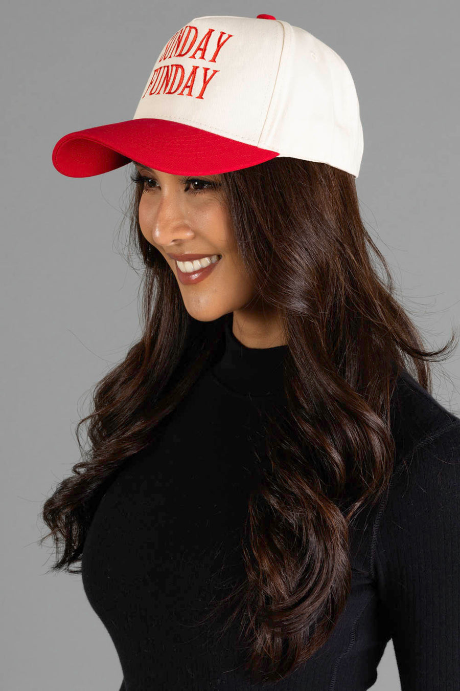 A woman with long dark hair, wearing a black top and the Panache Apparel Collection Sunday Funday Two-Toned Trucker Hat—a white-and-red adjustable snapback baseball cap featuring “SUNDAY FUNDAY” in red letters—smiles and faces left against a gray background.