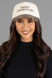 Smiling woman with long dark hair wears the Panache Apparel Collection Makin’ Cowboys Cry Ivory & Leopard Two-Toned Trucker Hat, paired with a black top and hoop earrings, standing against a plain gray background.