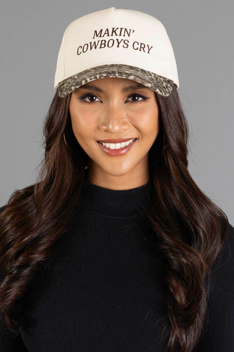 Smiling woman with long dark hair wears the Panache Apparel Collection Makin’ Cowboys Cry Ivory & Leopard Two-Toned Trucker Hat, paired with a black top and hoop earrings, standing against a plain gray background.