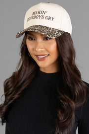 A woman with long dark hair is smiling and dressed in a black top, wearing the Panache Apparel Collection Makin’ Cowboys Cry Ivory & Leopard Two-Toned Trucker Hat, which features a cream base, leopard-print brim, and “MAKIN’ COWBOYS CRY” embroidered on the front.