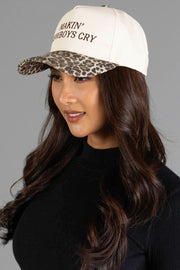 A woman with long brown hair is wearing the Panache Apparel Collection’s “Makin’ Cowboys Cry” Ivory & Leopard Two-Toned Trucker Hat, which features a cream color with leopard print bill and embroidered text, paired with a black top against a plain gray background.