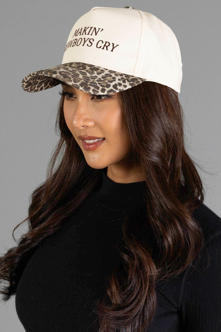 A woman with long brown hair is wearing the Panache Apparel Collection’s “Makin’ Cowboys Cry” Ivory & Leopard Two-Toned Trucker Hat, which features a cream color with leopard print bill and embroidered text, paired with a black top against a plain gray background.