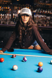 A woman wearing the Panache Apparel Collection Howdy Ivory & Leopard Two-Toned Trucker Hat and a brown shirt leans over a pool table surrounded by pool balls in a dimly lit bar with shelves of bottles in the background.