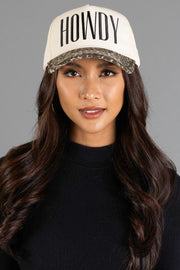 A woman with long dark hair is pictured against a plain gray background, wearing the Panache Apparel Collection Howdy Ivory & Leopard Two-Toned Trucker Hat—a cream-colored cap featuring bold black “HOWDY” lettering—paired with a black top and large hoop earrings.