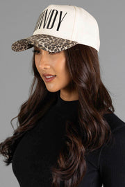 A woman with long brown hair is wearing the Panache Apparel Collection’s Howdy Ivory & Leopard Two-Toned Trucker Hat—a white cap featuring “Howdy” lettering and a leopard print brim—paired with a black ribbed top, photographed against a gray background.