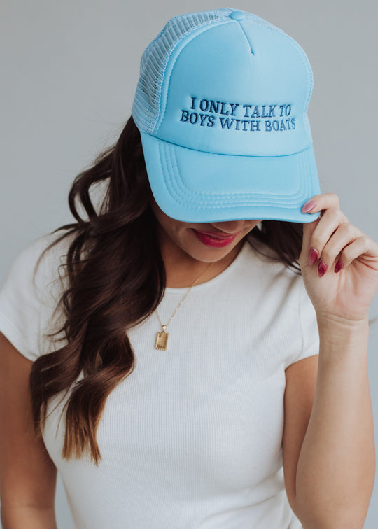 A woman wearing the panacheaccessories Boys With Boats Trucker Hat, styled with a white short-sleeve top and gold necklace, tilts her head down while holding the adjustable snapback’s brim.