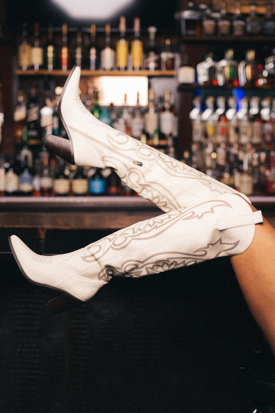 A person wearing Panache Apparel Collection’s Ivory Embroidered Western Boots rests their legs on a bar counter, with shelves of liquor bottles visible in the background.