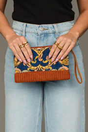 A person wearing blue jeans and a black top holds the Panache Apparel Collection’s Rust & Multicolored Patterned Wristlet, featuring vibrant orange and blue designs. Multiple gold rings adorn their fingers, and a wrist strap is attached to the wristlet.