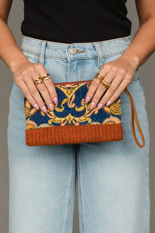 A person wearing blue jeans and a black top holds the Panache Apparel Collection’s Rust & Multicolored Patterned Wristlet, featuring vibrant orange and blue designs. Multiple gold rings adorn their fingers, and a wrist strap is attached to the wristlet.