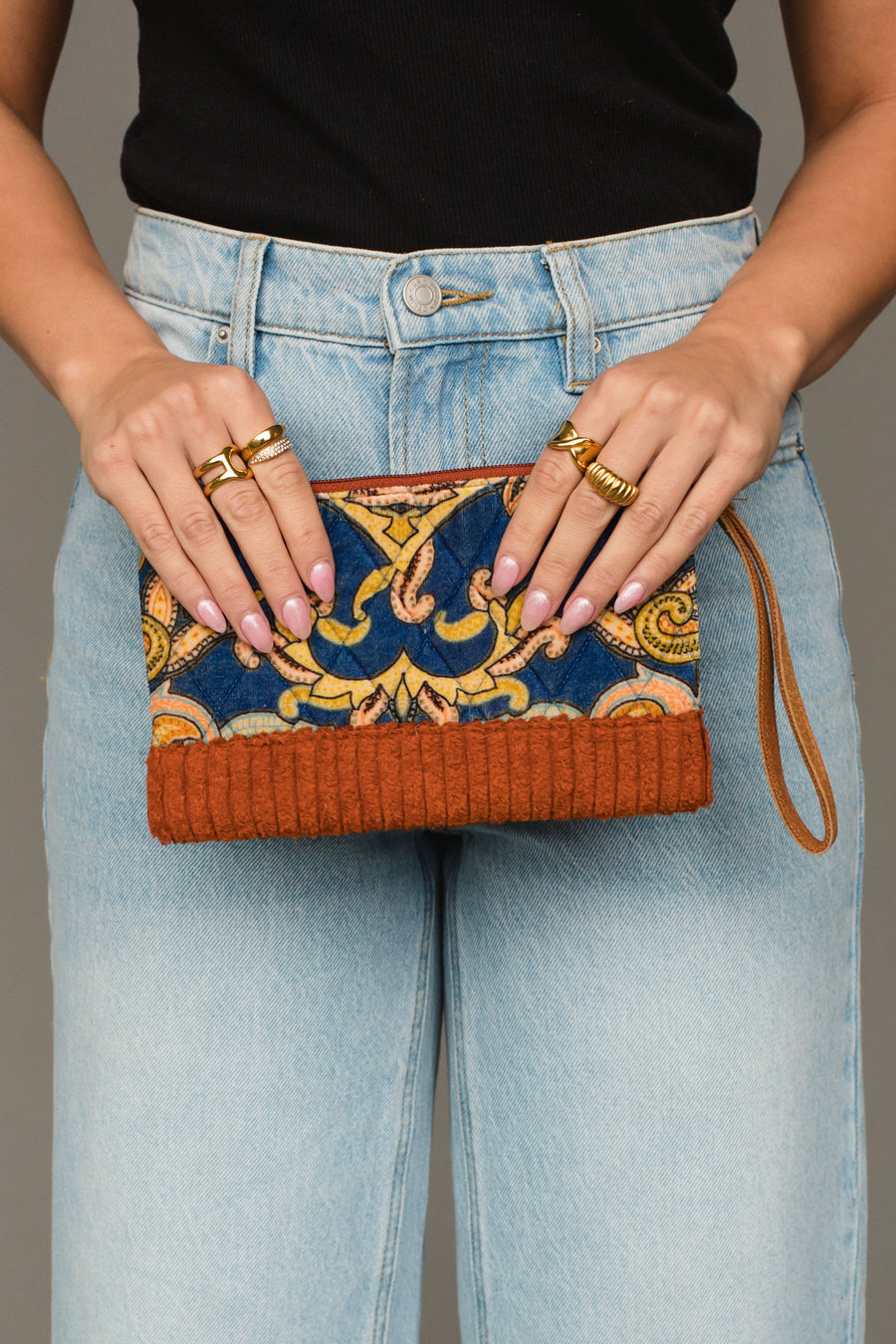 A person wearing blue jeans and a black top holds the Panache Apparel Collection’s Rust & Multicolored Patterned Wristlet, featuring vibrant orange and blue designs. Multiple gold rings adorn their fingers, and a wrist strap is attached to the wristlet.