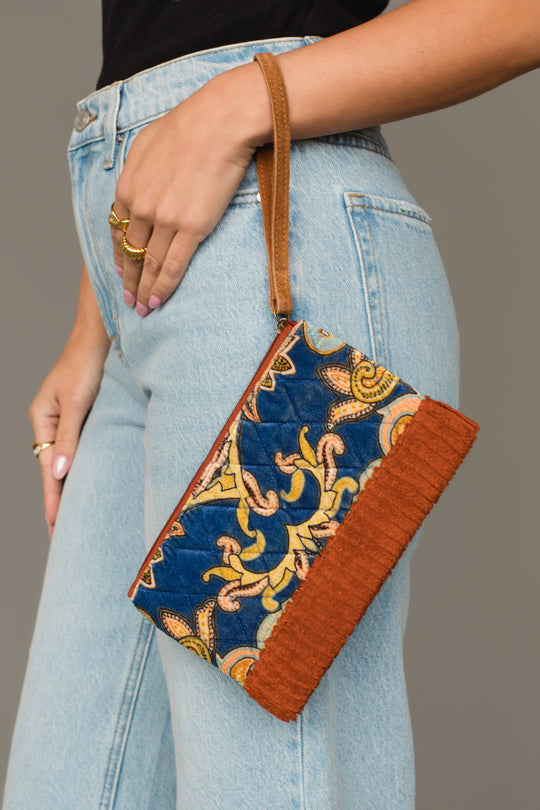 A person in light blue jeans holds the Panache Apparel Collection Rust & Multicolored Patterned Wristlet, featuring a brown strap, orange fabric border, and an intricate blue, yellow, and orange patterned design.