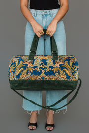 A person in light blue jeans and a black top holds the Panache Apparel Collection Green & Multicolored Patterned Duffel, featuring a colorful floral tapestry design and green corduroy straps. They wear black open-toe sandals, standing against a gray background.