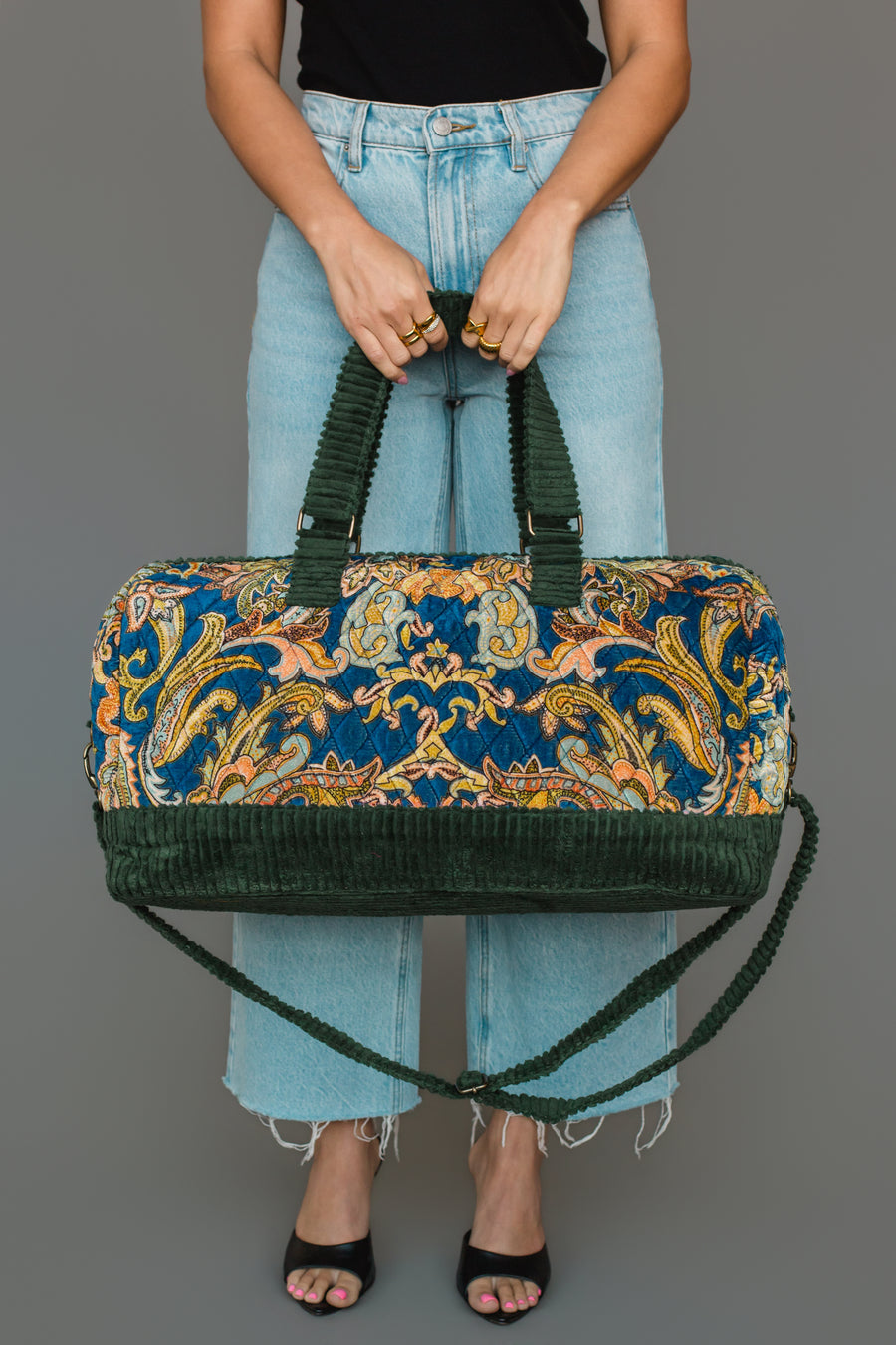 A person in light blue jeans and a black top holds the Panache Apparel Collection Green & Multicolored Patterned Duffel, featuring a colorful floral tapestry design and green corduroy straps. They wear black open-toe sandals, standing against a gray background.