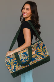 A woman smiles as she holds the Green & Multicolored Patterned Duffel by Panache Apparel Collection, featuring green handles and trim. She is dressed in a black shirt and light blue jeans, standing against a neutral background.