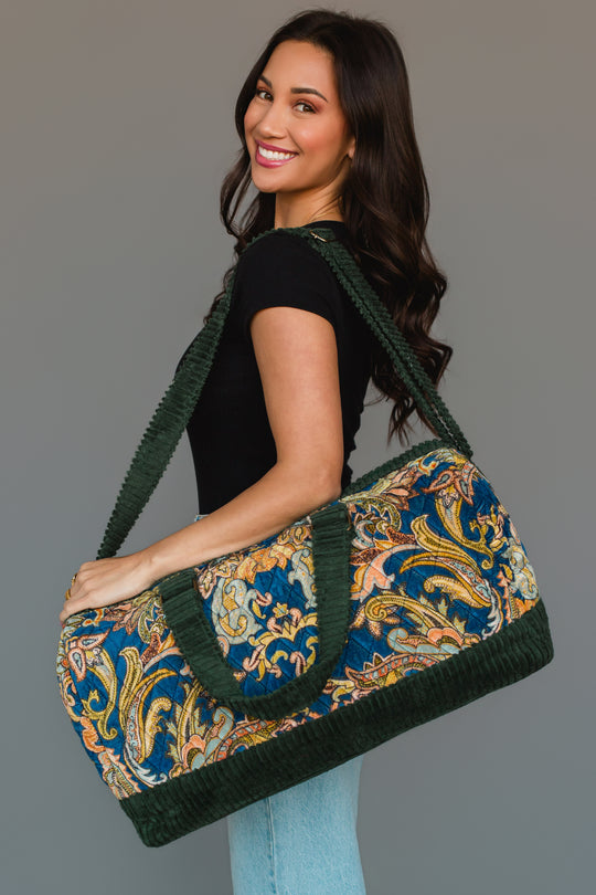 A woman smiles as she holds the Green & Multicolored Patterned Duffel by Panache Apparel Collection, featuring green handles and trim. She is dressed in a black shirt and light blue jeans, standing against a neutral background.