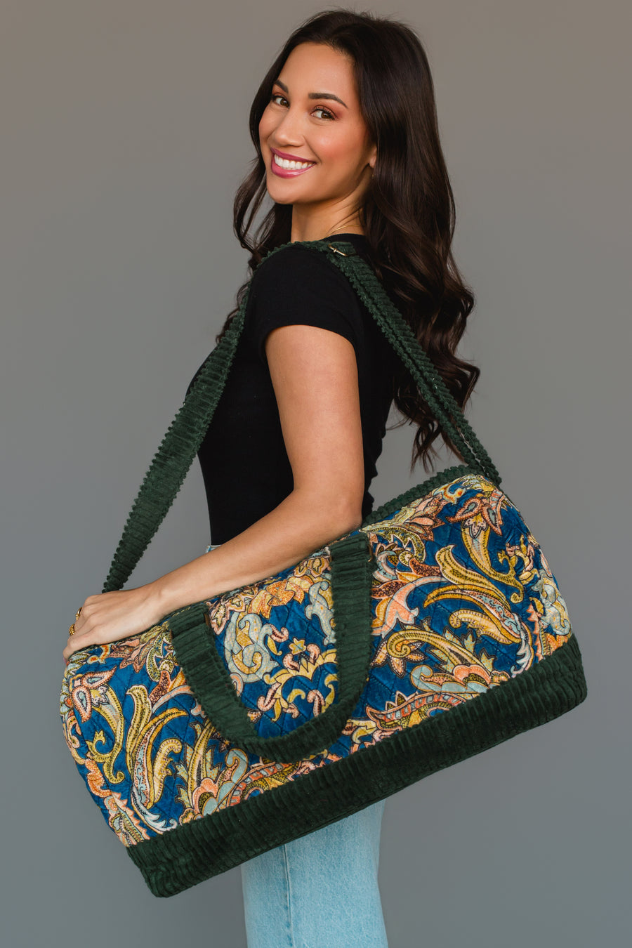 A woman smiles as she holds the Green & Multicolored Patterned Duffel by Panache Apparel Collection, featuring green handles and trim. She is dressed in a black shirt and light blue jeans, standing against a neutral background.