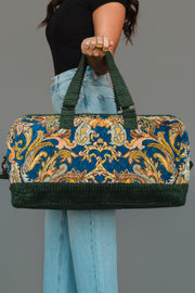 A person in a black shirt and blue jeans holds the Panache Apparel Collection Green & Multicolored Patterned Duffel, featuring green handles and trim, against a plain gray background.