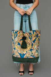 A person wearing light blue jeans and black sandals holds the Panache Apparel Collection Green & Multicolored Patterned Tote, which features vibrant paisley patterns, green braided handles, and a green tassel, standing against a plain gray background.