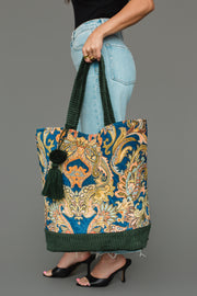 A woman carries the Panache Apparel Collection Green & Multicolored Patterned Tote, featuring a blue paisley print, green corduroy handles and base, and a dark green tassel. She pairs it with light jeans and black heels.