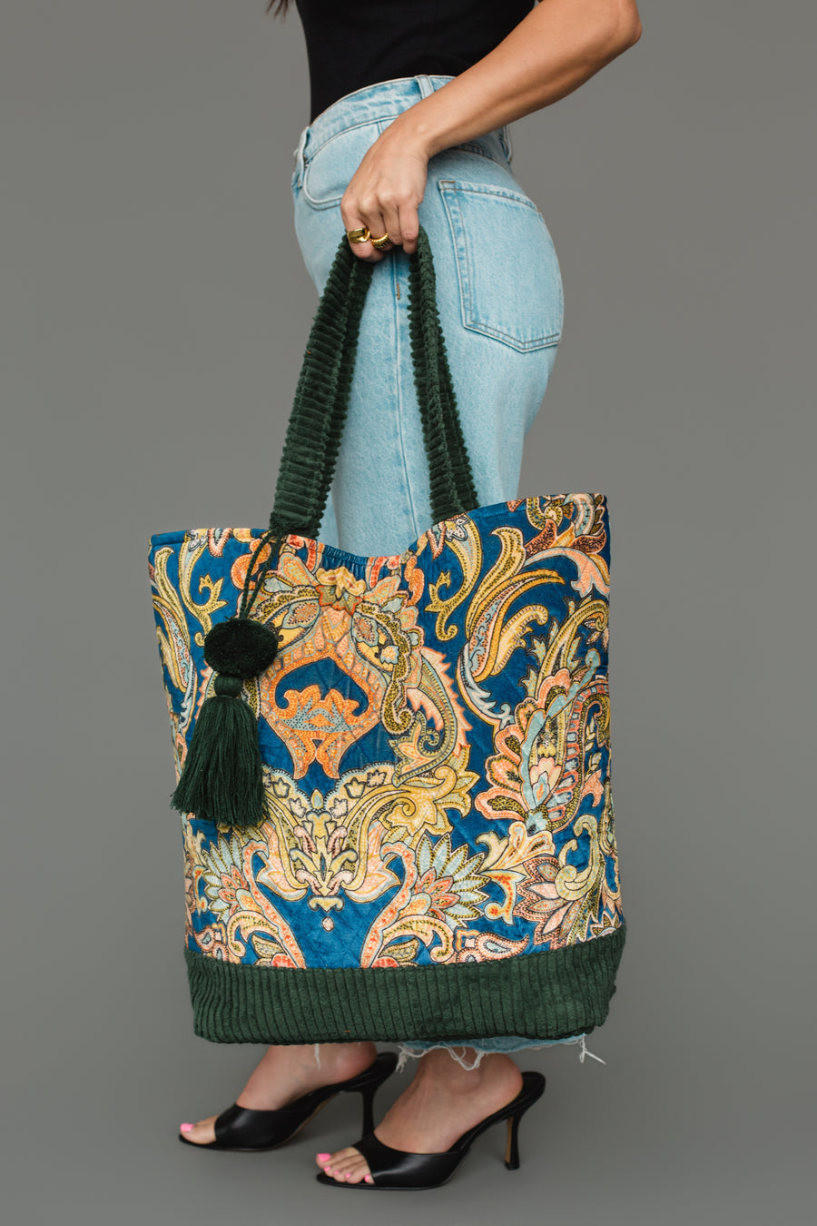 A woman carries the Panache Apparel Collection Green & Multicolored Patterned Tote, featuring a blue paisley print, green corduroy handles and base, and a dark green tassel. She pairs it with light jeans and black heels.