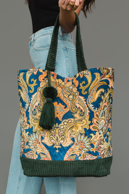 A person holding the Panache Apparel Collection Green & Multicolored Patterned Tote, featuring green handles, a large green tassel, and dark green corduroy trim, styled with light blue jeans and a black top.