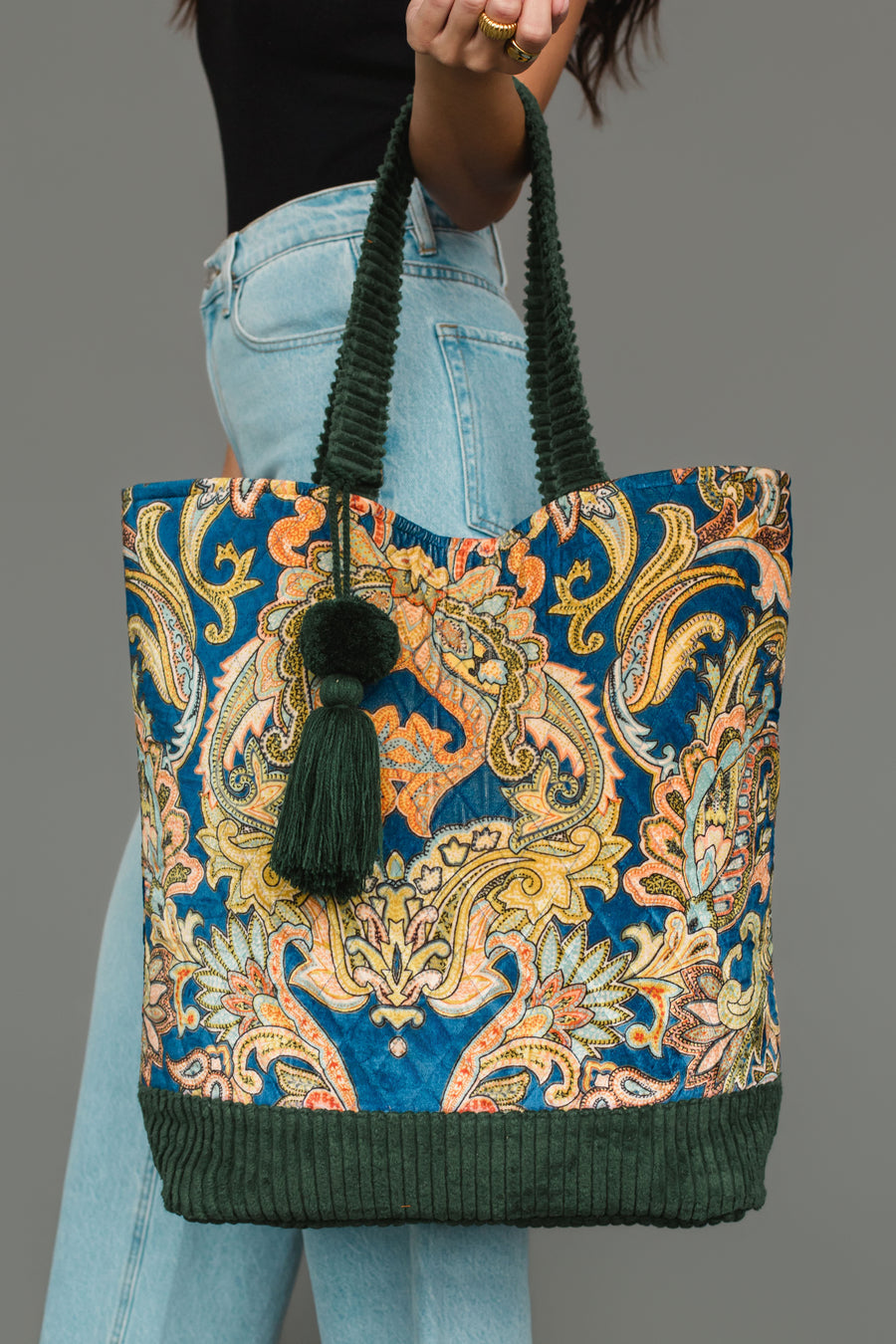 A person holding the Panache Apparel Collection Green & Multicolored Patterned Tote, featuring green handles, a large green tassel, and dark green corduroy trim, styled with light blue jeans and a black top.