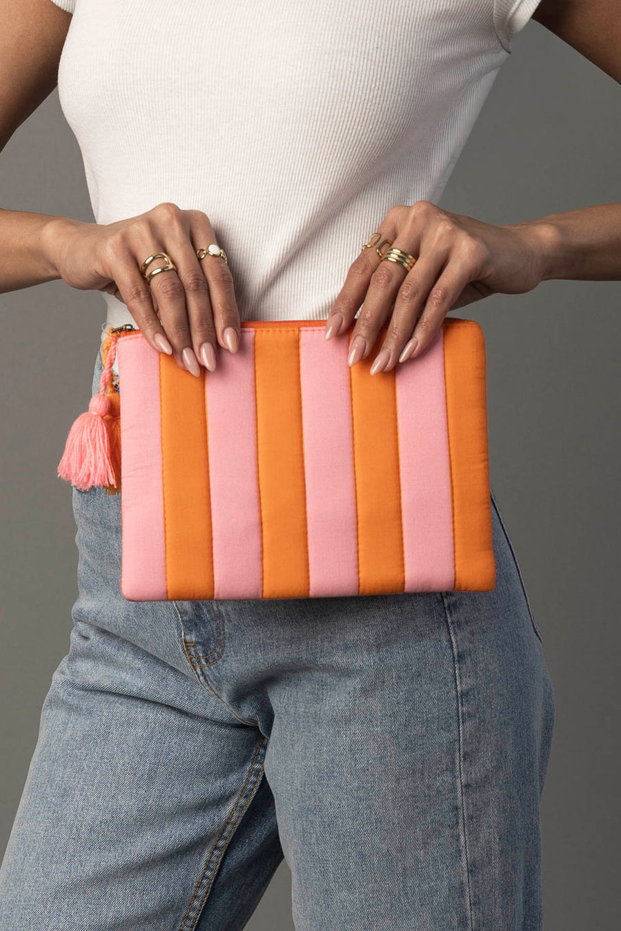 A person wearing a white top and blue jeans holds the Panache Apparel Collection Light Pink & Orange Striped Clutch, which features a tassel zipper, while displaying neatly manicured nails and several rings.