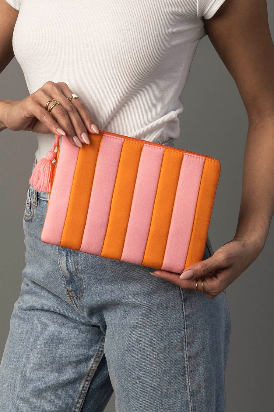 A person wearing a white shirt and blue jeans holds the Panache Apparel Collection Light Pink & Orange Striped Clutch, which features vertical orange and pink stripes and a tassel on the zipper.