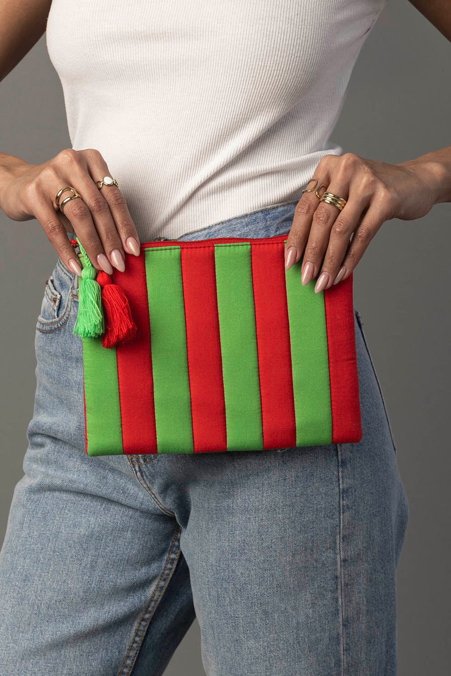 A person in a white tank top and blue jeans holds the Panache Apparel Collection Pink & Green Striped Clutch with tassels, showing off rings on their fingers.
