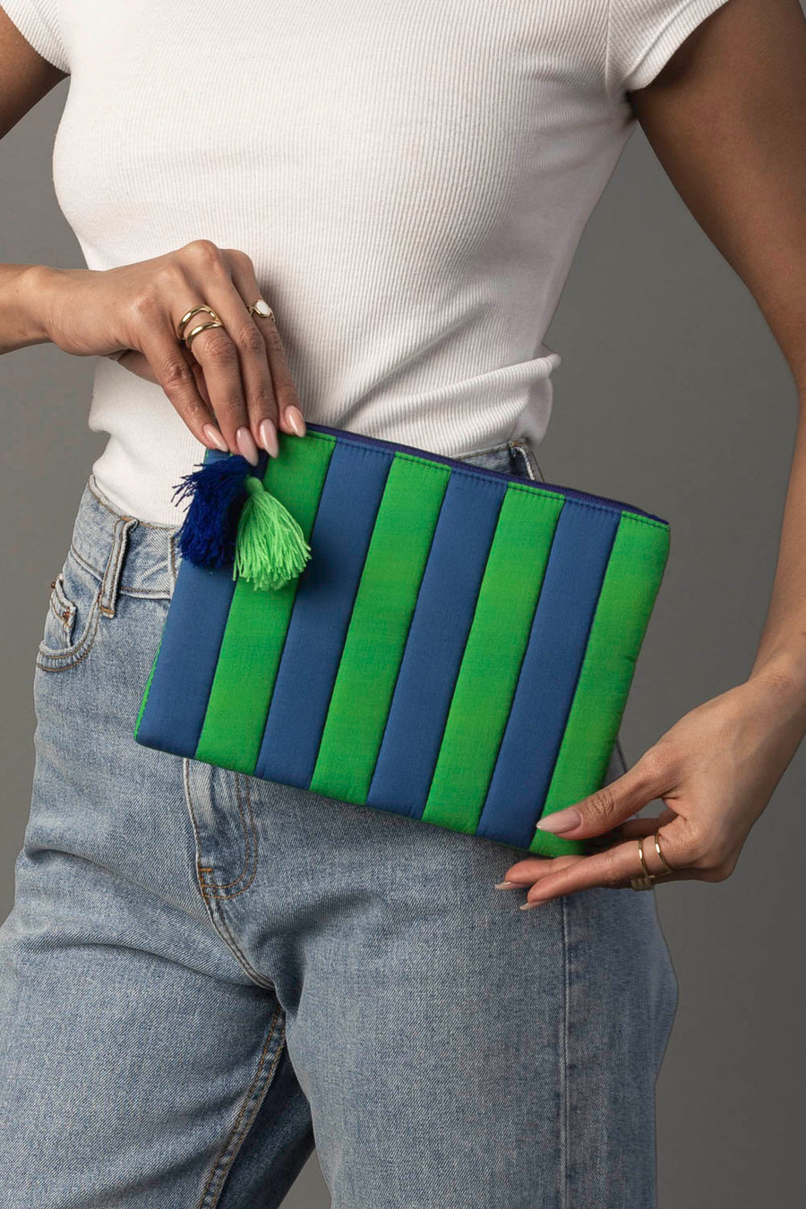 A person in a white top and jeans holds the Panache Apparel Collection Blue & Green Striped Clutch, which features a green and blue tassel.