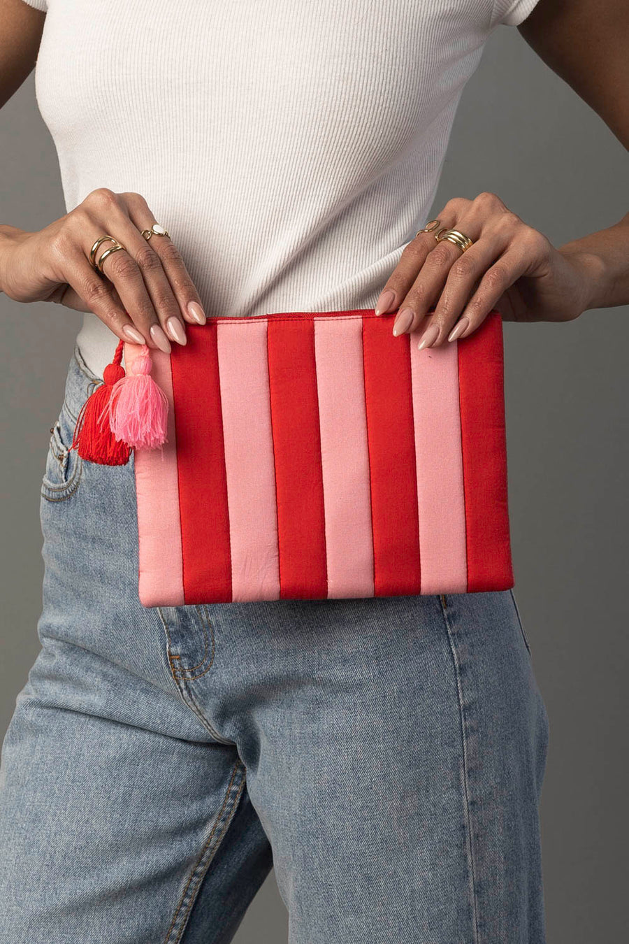 A person in a white ribbed top and jeans holds the Panache Apparel Collection Red & Light Pink Striped Clutch, featuring a pink and red tassel, against a plain gray background.
