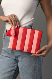A person in a white sleeveless top and blue jeans holds the Panache Apparel Collection Red & Light Pink Striped Clutch, featuring a red and pink tassel.