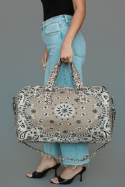 A woman in light blue jeans and black heels holds the Panache Apparel Collection Tan Bandana Duffel, featuring a bold paisley pattern, as she stands against a plain grey background.