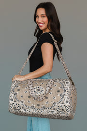 A woman in a black top and jeans smiles while holding the Panache Apparel Collection’s Tan Bandana Duffel, a large patterned beige bag with two handles and a shoulder strap, against a plain background.
